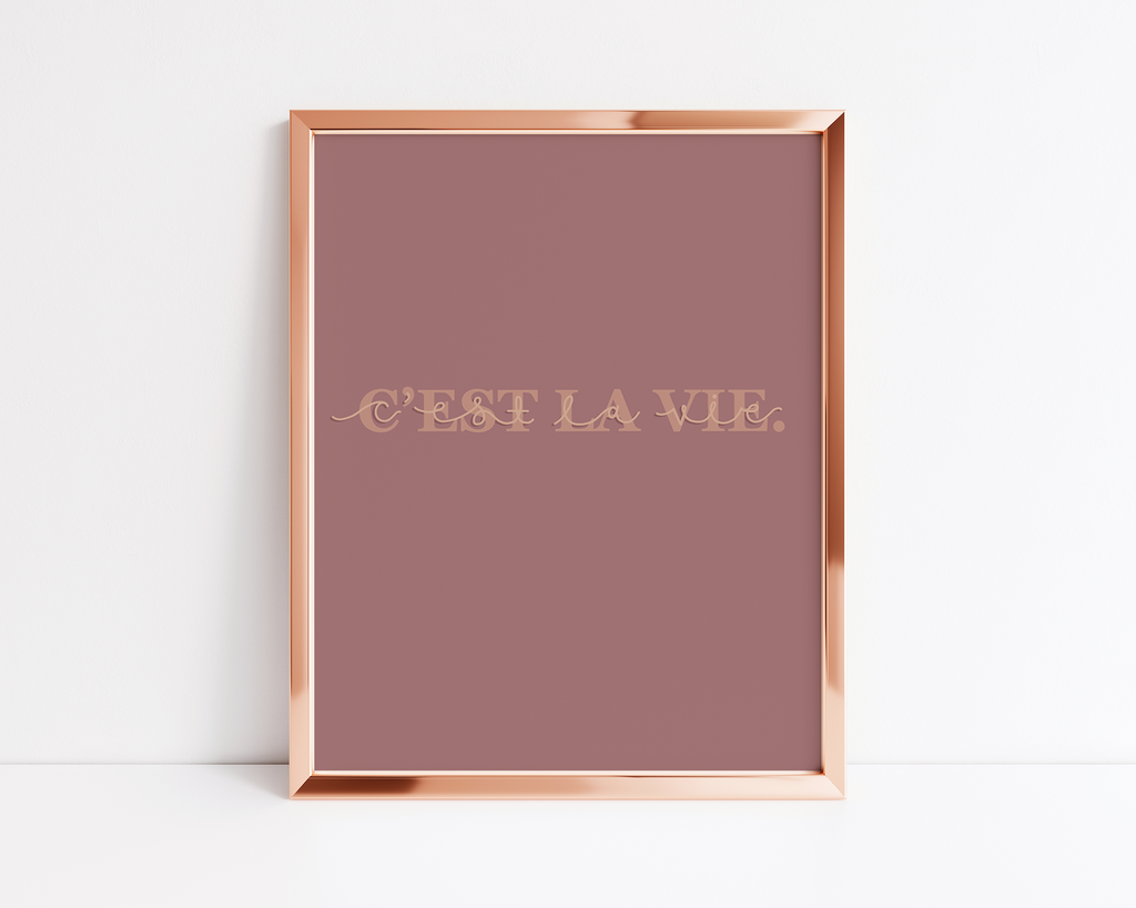 A print in a rose gold frame with a dark blush background and the phrase "C'EST LA VIE" in serif block letters with cursive overlay 